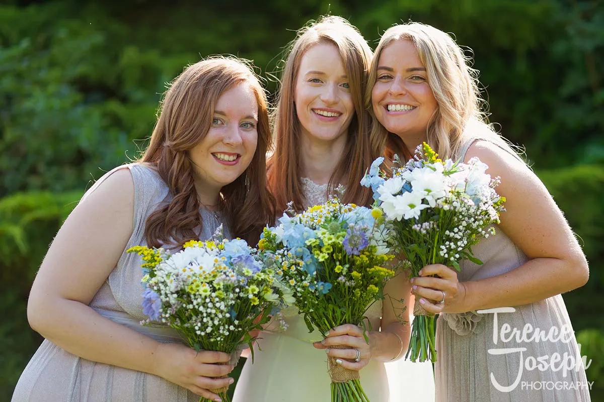 36_Moreteyne_Manor_Wedding_Breakfast Three women in light dresses smile and hold bouquets of flowers, standing outdoors in front of leafy greenery. Bright sunlight highlights their faces as they enjoy the moment together, perfectly captured by a Moreteyne Manor wedding photographer.