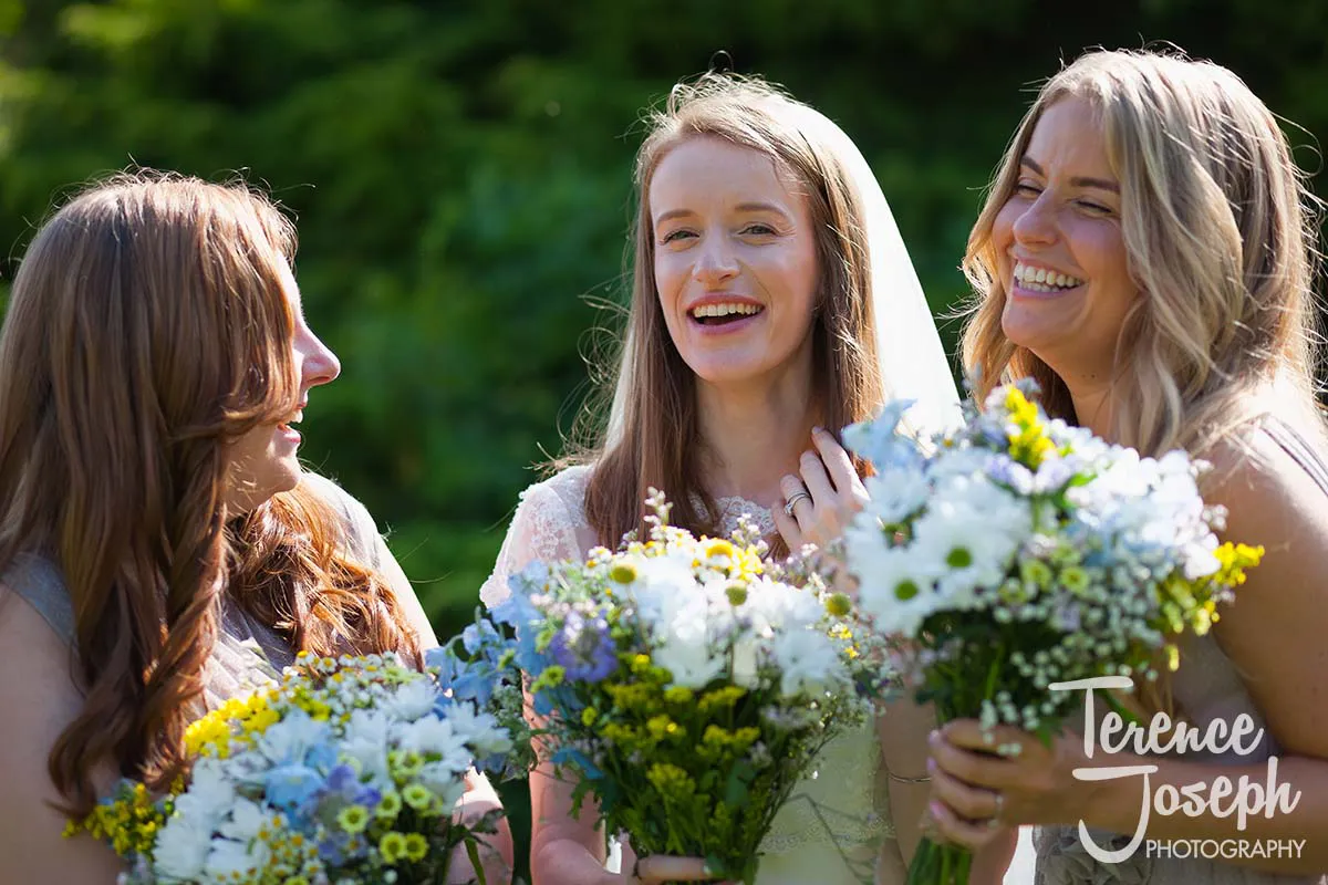 37_Moreteyne_Manor_Wedding_Breakfast Three women in bright dresses share a joyful moment outdoors at Moreteyne Manor. The bride, in the center, holds a bouquet of white and yellow flowers, flanked by bridesmaids with similar bouquets. They are bathed in sunlight with greenery as a backdrop, captured perfectly by the wedding photographer.