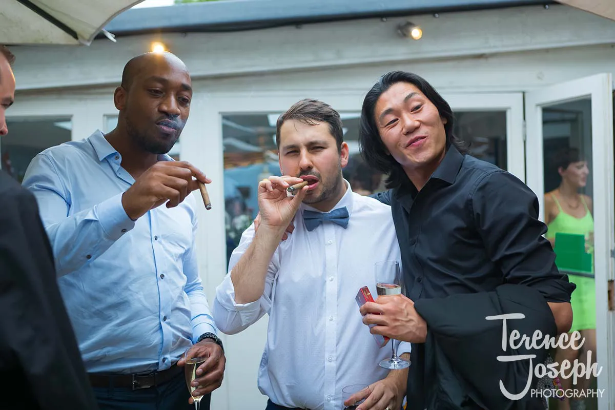 38_County_Arms_Wandsworth_Wedding_Breakfast_Photos Three men in formal attire holding cigars and drinks gather outdoors, seemingly at a County Arms Wandsworth wedding. With a white building and mingling guests in the background, the mood is relaxed and festive.