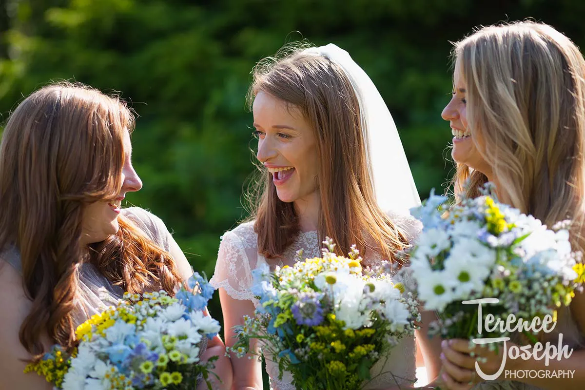38_Moreteyne_Manor_Wedding_Breakfast Three women, dressed in light outfits, share a joyful moment outdoors, holding bouquets of white, blue, and yellow flowers. The woman in the center wears a veil, hinting at a bridal setting—a perfect scene for any Moreteyne Manor wedding photographer. Lush greenery forms the backdrop.