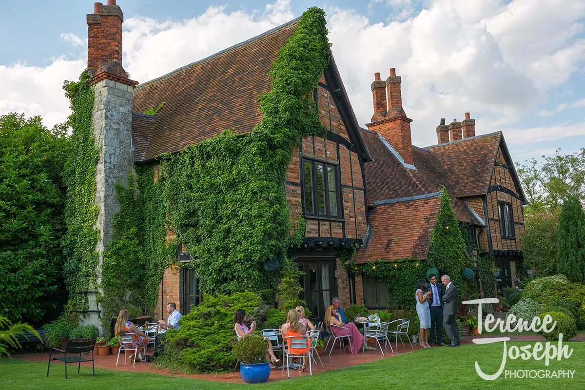 39_Moreteyne_Manor_Wedding_Breakfast A picturesque brick house covered in ivy, with a sloped roof and multiple chimneys. People are seated and standing around tables in a lush garden, enjoying the outdoor setting. The Moreteyne Manor Wedding Photographer captures the charm as partly cloudy skies add to the tranquil atmosphere.