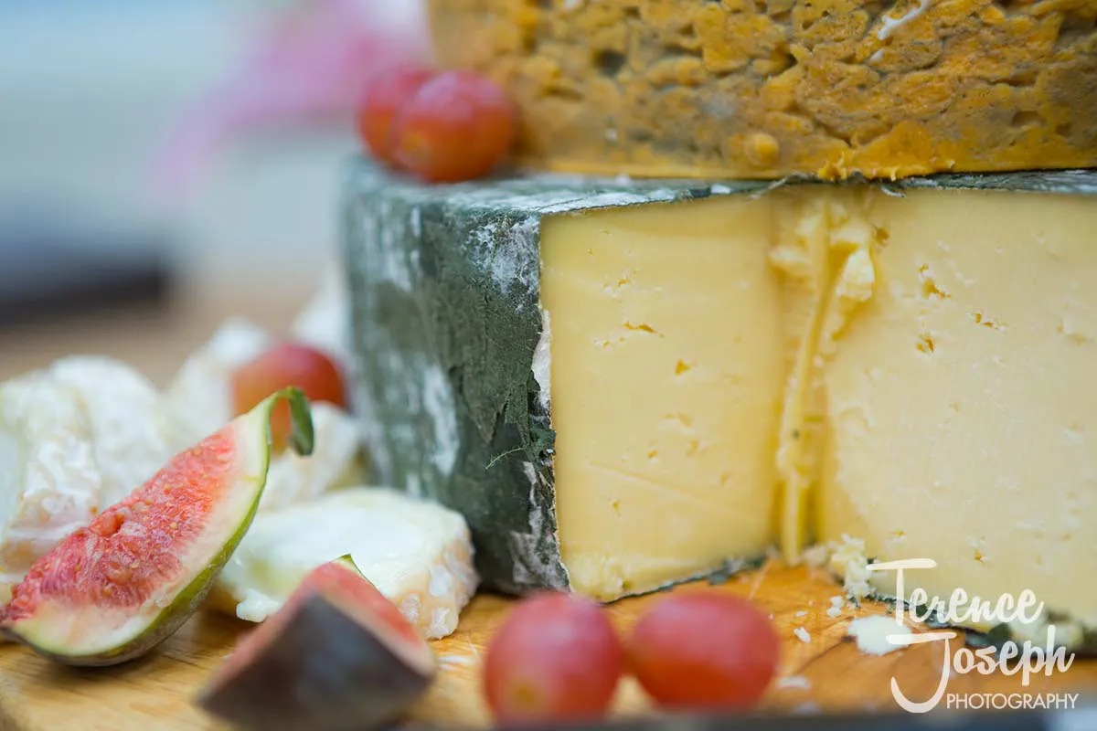 40_County_Arms_Wandsworth_Wedding_Breakfast_Photos Close-up of a cheese wheel on a wooden board, with a wedge cut out. The cheese, perfect for a County Arms Wandsworth wedding, features a textured rind and creamy interior. Surrounding it are red grapes, figs, and soft white cheese.