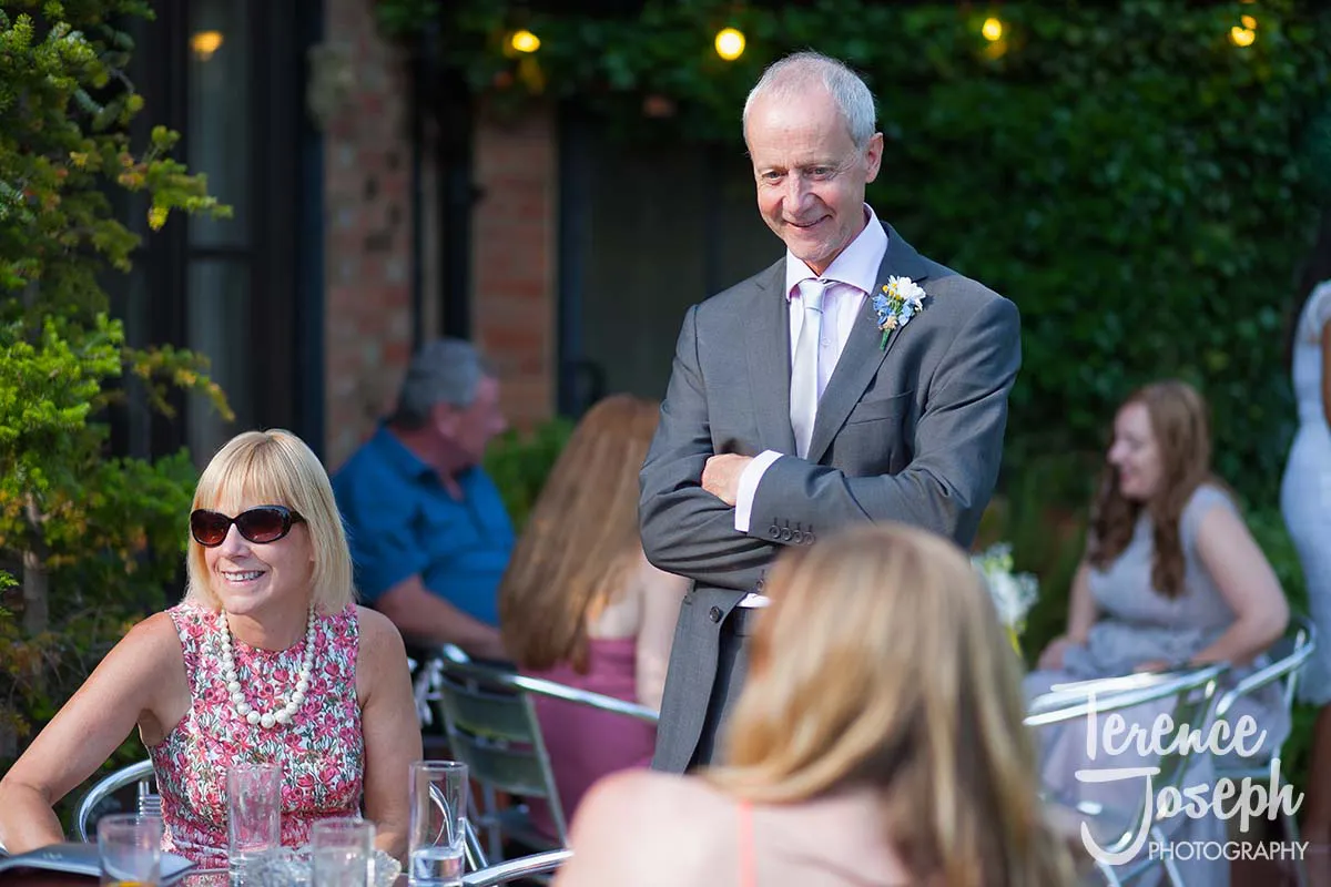 40_Moreteyne_Manor_Wedding_Breakfast A man in a suit with a boutonnière stands smiling, arms crossed, beside a seated woman in sunglasses and a floral dress at an outdoor gathering. Capturing this moment as their Moreteyne Manor wedding photographer, I loved the lively backdrop of people and greenery.