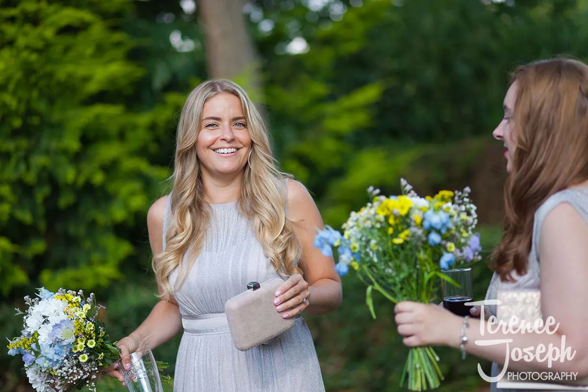41_Moreteyne_Manor_Wedding_Breakfast In a picturesque garden setting, two women exude joy. One, with long blonde hair and a light sleeveless dress, smiles while holding a bouquet and glass. Her companion, with long light brown hair, mirrors her delight. Such moments are beautifully captured by a Moreteyne Manor Wedding Photographer amidst lush greenery.