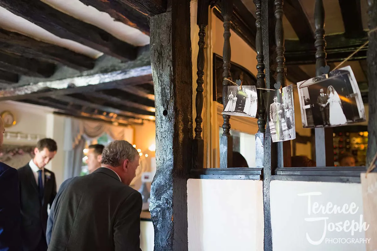42_Moreteyne_Manor_Wedding_Breakfast A few people in formal attire engage in conversation inside a rustic room with wooden beams. Black and white wedding photos hang on a string, attached with clips. A sign reads "Moreteyne Manor Wedding Photographer" in the bottom right corner.