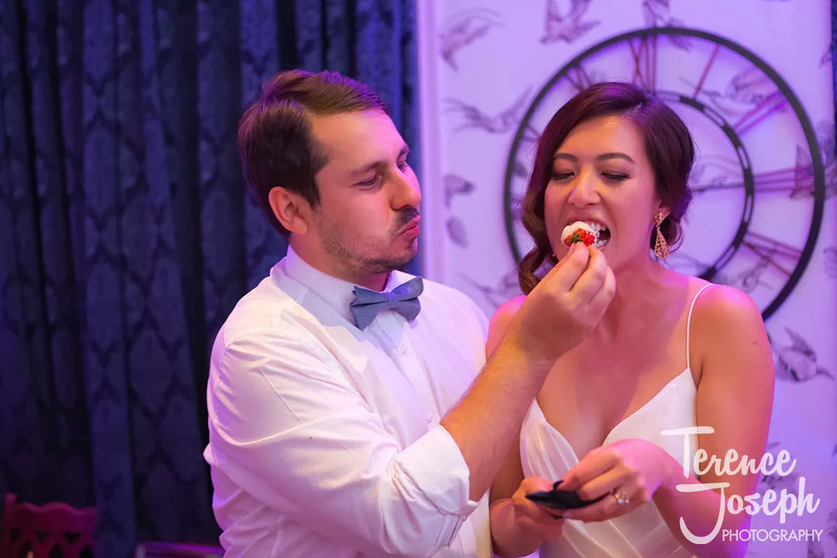 43_County_Arms_Wandsworth_Evening_Wedding_Recption At a charming County Arms Wandsworth wedding, the groom, sporting a bow tie, lovingly feeds cake to his bride in her white dress. They stand indoors with a decorative clock behind them, the photo watermarked "Terence Joseph Photography.