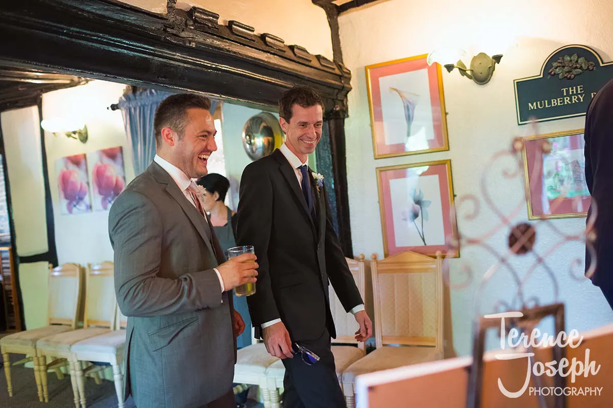 43_Moreteyne_Manor_Wedding_Breakfast Two men in suits are smiling and walking indoors at an event venue, perhaps captured by a Moreteyne Manor Wedding Photographer. One holds a drink while the other sports a boutonniere. Framed artwork decorates the walls, and a sign reads "The Mulberry," with tables and chairs visible in the background.