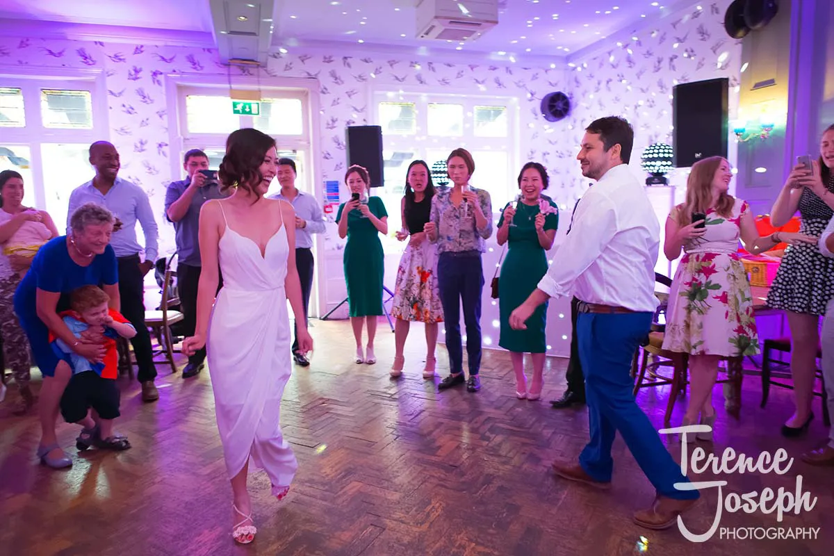 44_County_Arms_Wandsworth_Evening_Wedding_Recption A joyful couple dances in the center of a vibrant County Arms Wandsworth wedding. Guests gather around, smiling, clapping, and taking photos as colorful lights illuminate the scene. A child is being held by an older guest on the left.