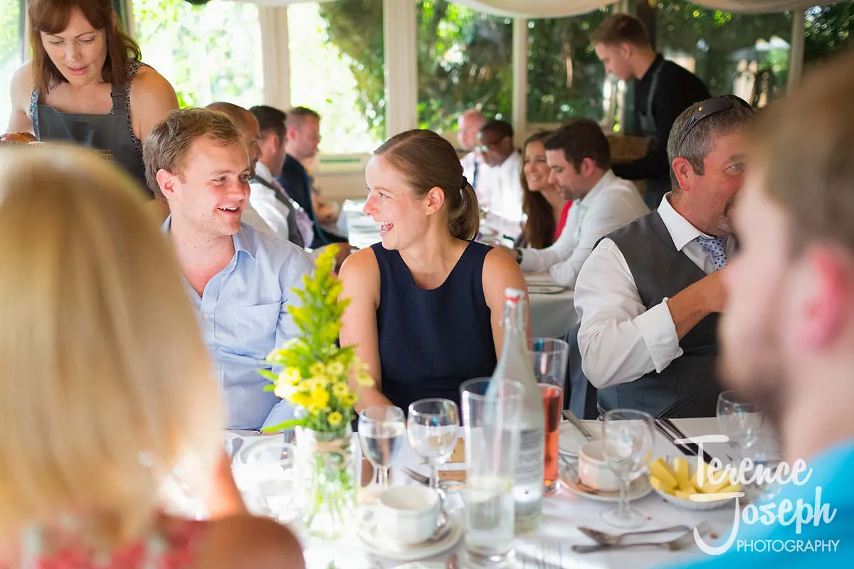 44_Moreteyne_Manor_Wedding_Breakfast At a social event, a group sits around an elegantly set dining table adorned with glassware and a vase of yellow flowers. Two people smile in lively conversation, capturing moments reminiscent of a Moreteyne Manor Wedding Photographer's enchanting touch.