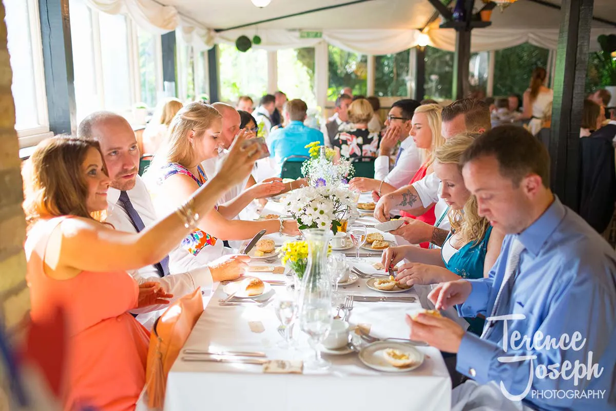 45_Moreteyne_Manor_Wedding_Breakfast People seated at a long, elegantly set dining table, enjoying a meal together. Sunlight streams through the windows of Moreteyne Manor, creating a warm atmosphere. The room is decorated with white drapes and floral centerpieces adorn the tables—an ideal scene for a wedding photographer.