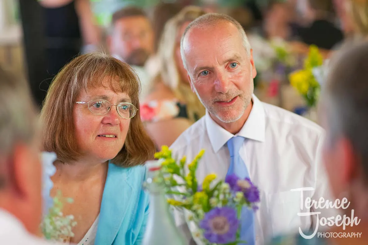 46_Moreteyne_Manor_Wedding_Breakfast At an event reminiscent of a Moreteyne Manor wedding, a man in a light blue tie and a woman in a matching blue outfit sit at a table adorned with floral centerpieces, engrossed in conversation. Other attendees form the backdrop, adding to this picturesque setting.