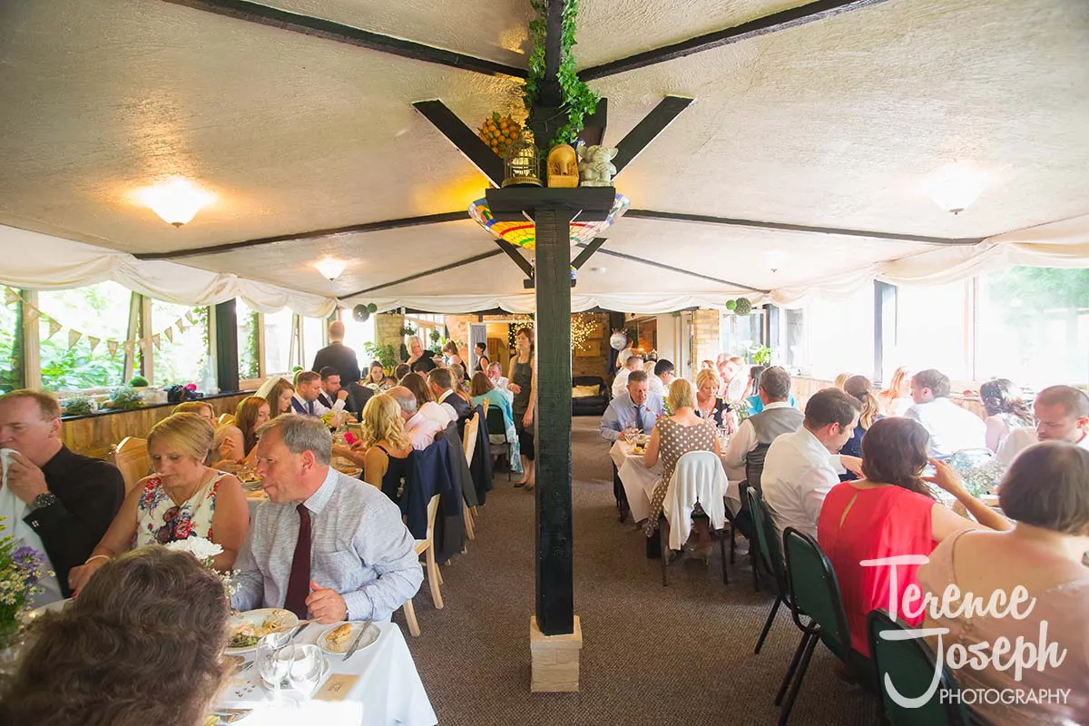 47_Moreteyne_Manor_Wedding_Breakfast At this elegant Moreteyne Manor wedding, guests gather at long tables, savoring exquisite dishes and lively conversation. The room is adorned with white drapes and greenery, while a black pillar with hanging bottles stands central. Large windows flood the space with natural light.