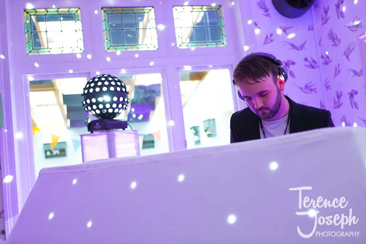 48_County_Arms_Wandsworth_Evening_Wedding_Recption At a County Arms Wandsworth wedding, a DJ wearing headphones stands behind a table draped in white cloth, illuminated by colorful disco lights. A mirror ball spins, casting shimmering reflections alongside the decorative windows in the background.