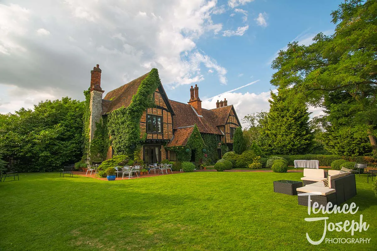 48_Moreteyne_Manor_Wedding_Breakfast A charming, ivy-covered cottage with a steep roof and tall chimneys stands gracefully amidst a lush garden. Outdoor seating with tables and chairs is arranged on the lawn under a partly cloudy sky, perfect for capturing timeless moments as Moreteyne Manor Wedding Photographer.