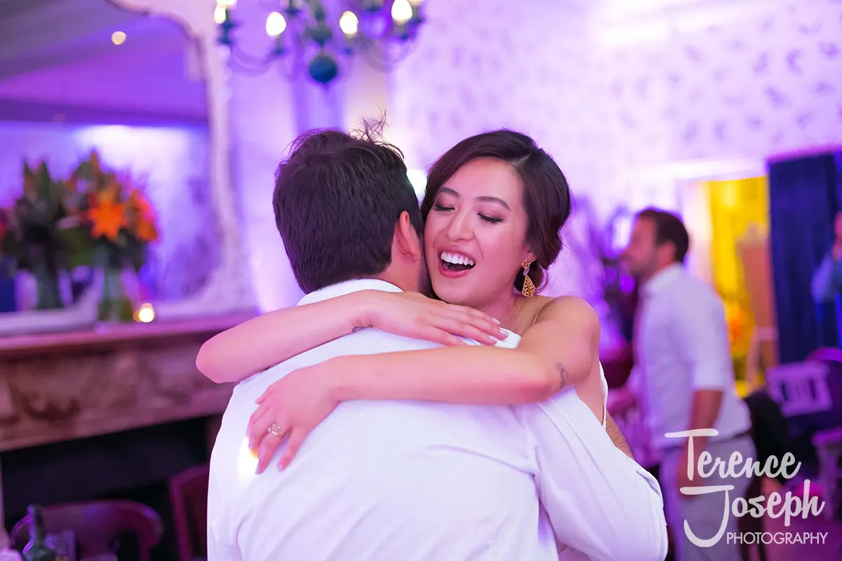 50_County_Arms_Wandsworth_Evening_Wedding_Recption A joyful woman embraces a man at the County Arms Wandsworth wedding reception, both dressed in formal attire. The background is softly lit with purple hues, featuring blurred partygoers and floral arrangements.