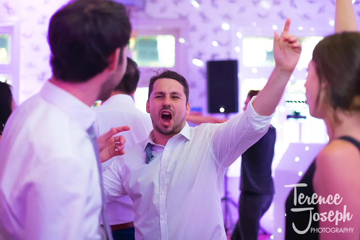 51_County_Arms_Wandsworth_Evening_Wedding_Recption A man in a white shirt is excitedly dancing and pointing upwards at a wedding party at the County Arms Wandsworth. He is surrounded by other people on the dance floor in a lively atmosphere, with colorful lights in the background.