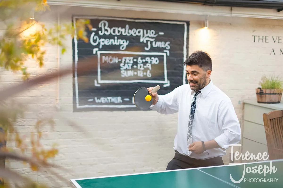 53_County_Arms_Wandsworth_Evening_Wedding_Recption A man in a white shirt and tie enjoys a lively game of table tennis outdoors, near the County Arms Wandsworth. Behind him, a chalkboard cheerfully displays the "Barbeque Opening Times," adding to the vibrant scene.
