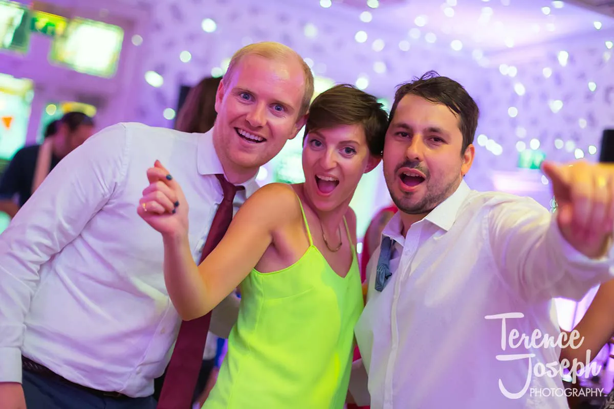 54_County_Arms_Wandsworth_Evening_Wedding_Recption Three people smiling and posing for the camera at a wedding party at County Arms Wandsworth, with one man holding a microphone. The background is colorful with soft, glowing lights, adding a festive touch to the joyful celebration.