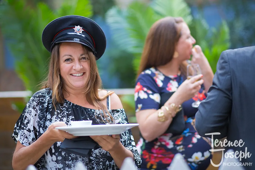 01_Bermondsey_Yard_Café_Wedding_Photographer_ 0001 A smiling woman in a police-style hat holds a tray with cups at a Bermondsey Yard Café Wedding, while people in the background talk and drink. The scene is bright and cheerful, with green plants behind them.