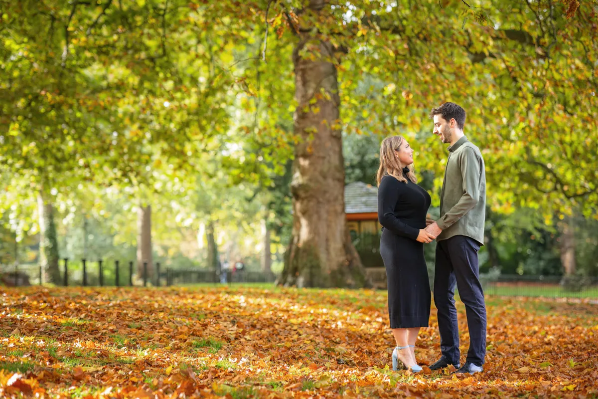A couple standing and holding hands in a park during their autumn engagement photo session, surrounded by fallen leaves, with trees displaying fall colors in the background.