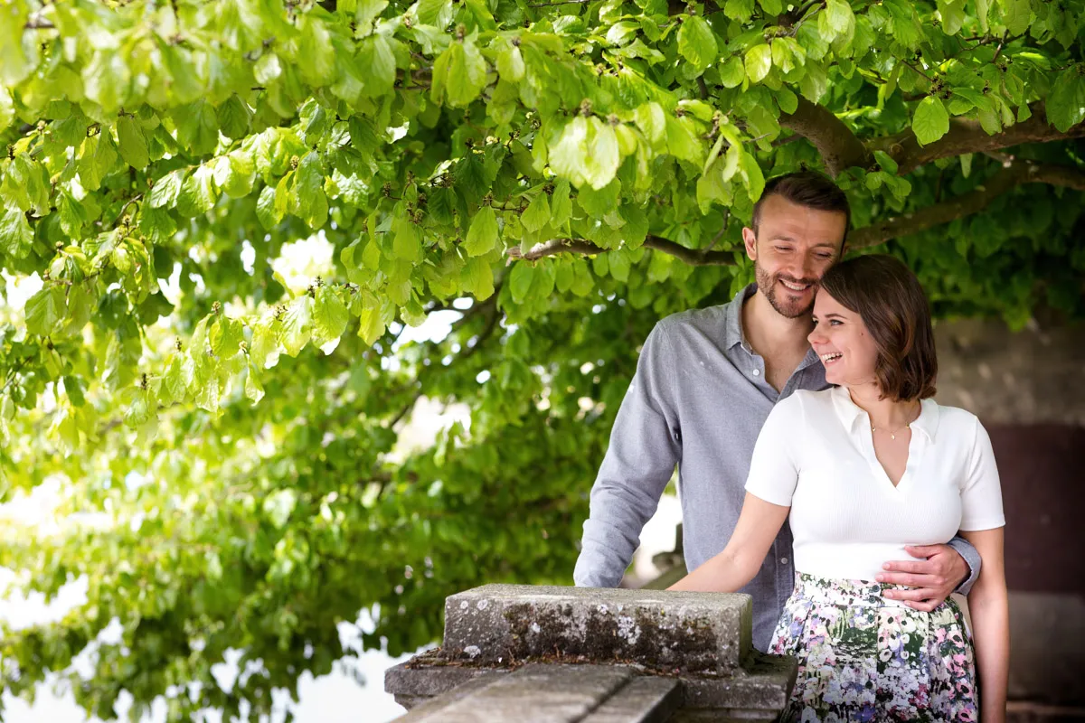 A couple stands affectionately under a lush green tree during their London engagement session. The woman, wearing a white top and floral skirt, smiles while leaning against a stone railing. The man, in a grey shirt, stands close behind her, smiling and holding her waist.