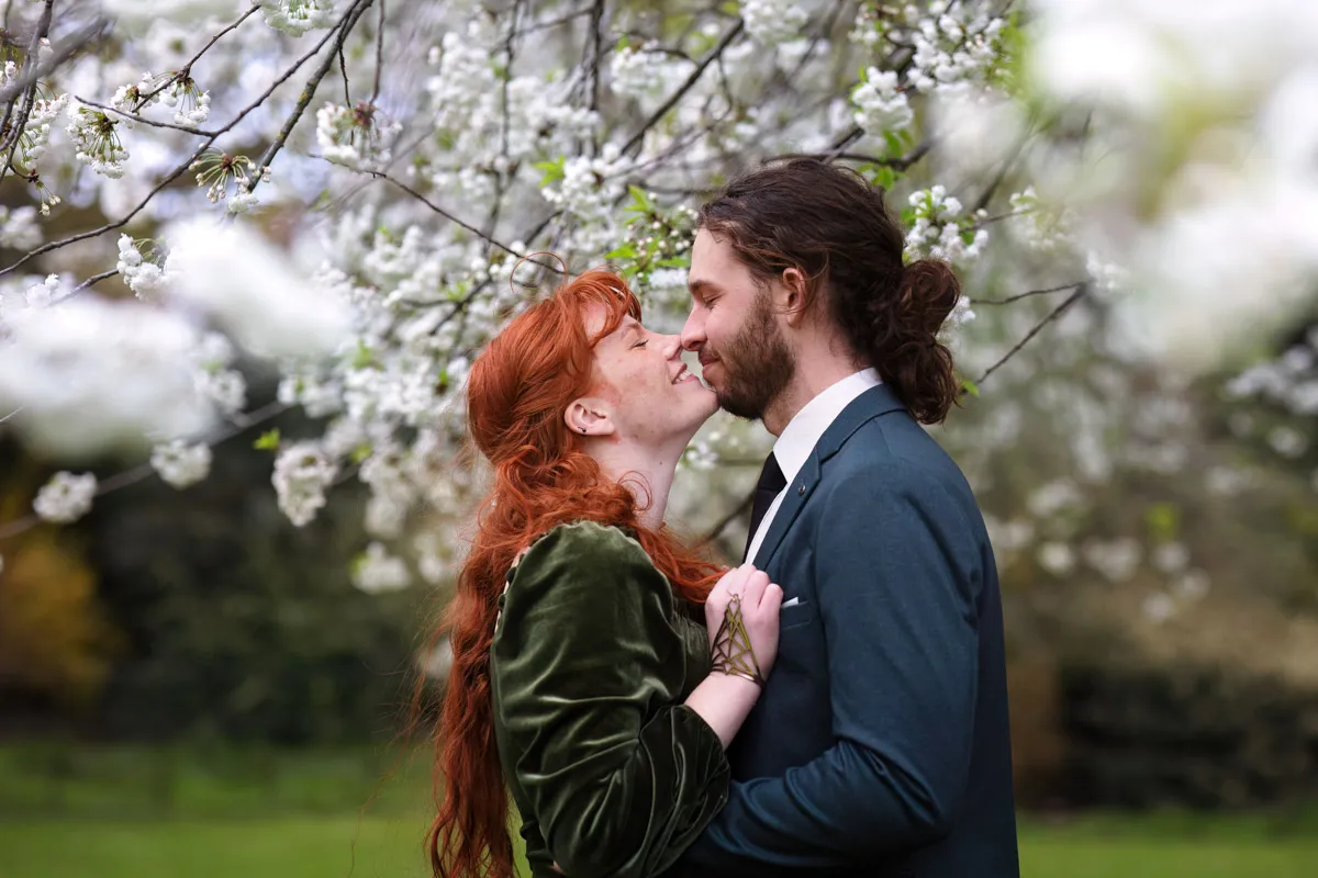 A couple embraces and shares a tender moment under a canopy of white blossoming trees during their engagement photoshoot, with the woman's red hair contrasting brightly against the lush green background.