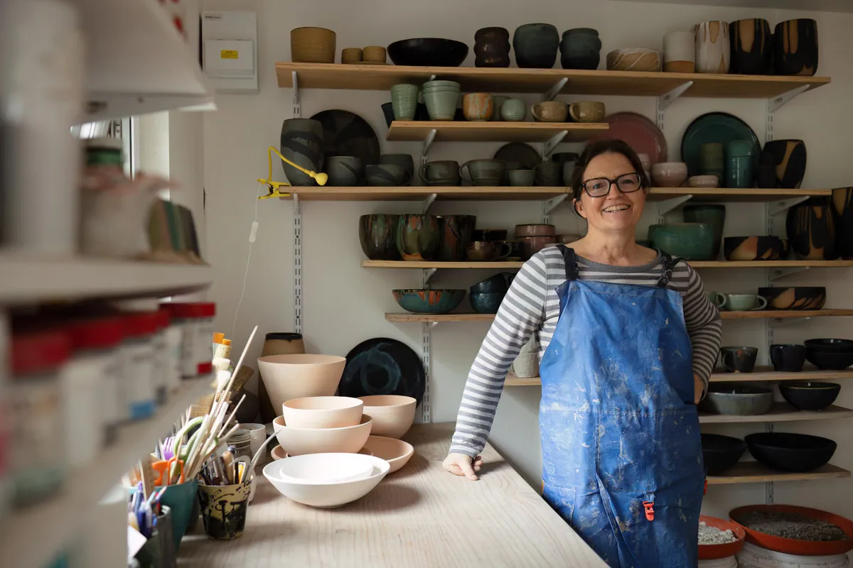 TJP_2024_04_25_0071 A potter in a blue apron stands proudly in her studio, surrounded by shelves filled with colorful ceramic bowls and plates. She smiles beside a worktable with clay tools and pottery in progress, perfectly
