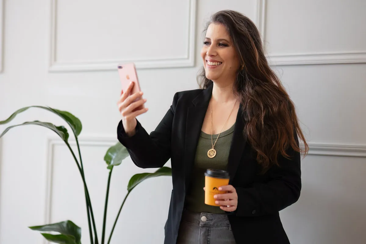 TJP_2024_04_26_0004 A smiling woman in a business casual outfit holds a smartphone and a coffee cup, taking a selfie in a well-lit room with elegant white paneling and a plant in the background for her wellness coach
