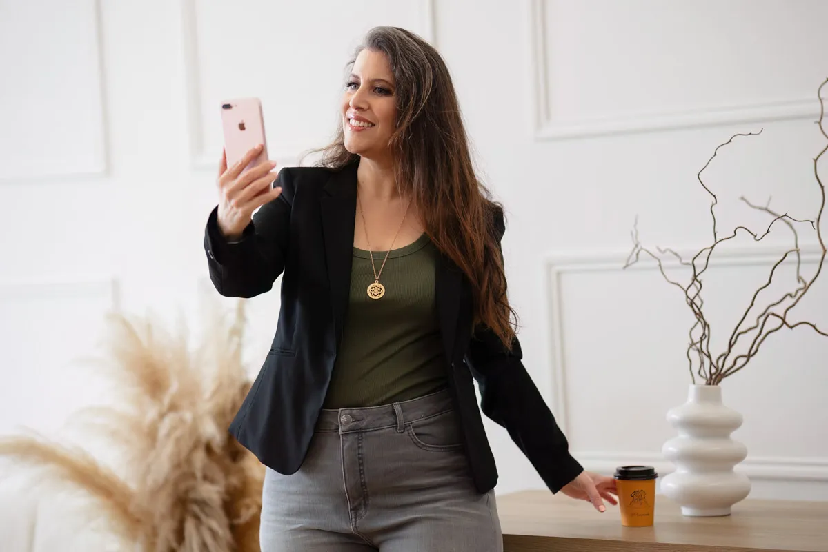 TJP_2024_04_26_0006 A woman with long hair takes a selfie with her phone, smiling and standing by a table with a coffee cup and decorative vases in a stylish room designed for brand photography.