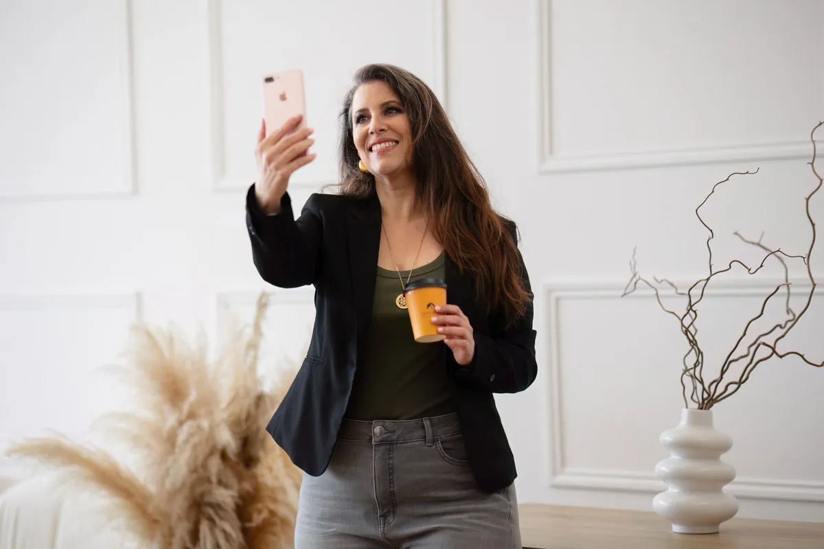 TJP_2024_04_26_0008 A wellness coach takes a selfie with her phone in one hand and holds a coffee cup in the other, smiling in a stylishly decorated room.