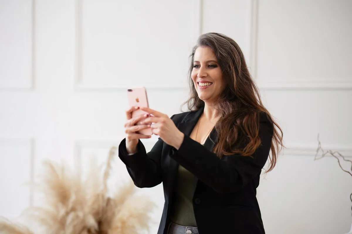 TJP_2024_04_26_0012 A woman with long hair smiles as she takes a photo using her smartphone in a bright, elegant room. She's dressed as a wellness coach in a black blazer and a green top.