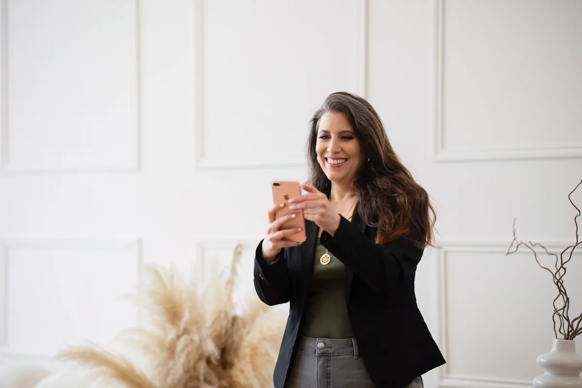 TJP_2024_04_26_0014 A smiling wellness coach in a black blazer and green top using her smartphone, possibly taking a selfie, in a softly lit room with elegant decor.