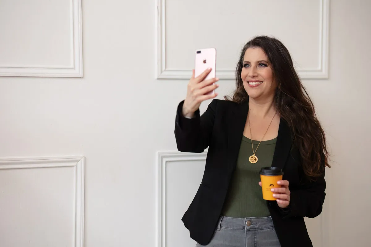 TJP_2024_04_26_0048 A wellness coach with long brown hair, wearing a black blazer and green top, takes a selfie with her phone while holding a coffee cup in her other hand, against a white paneled wall.