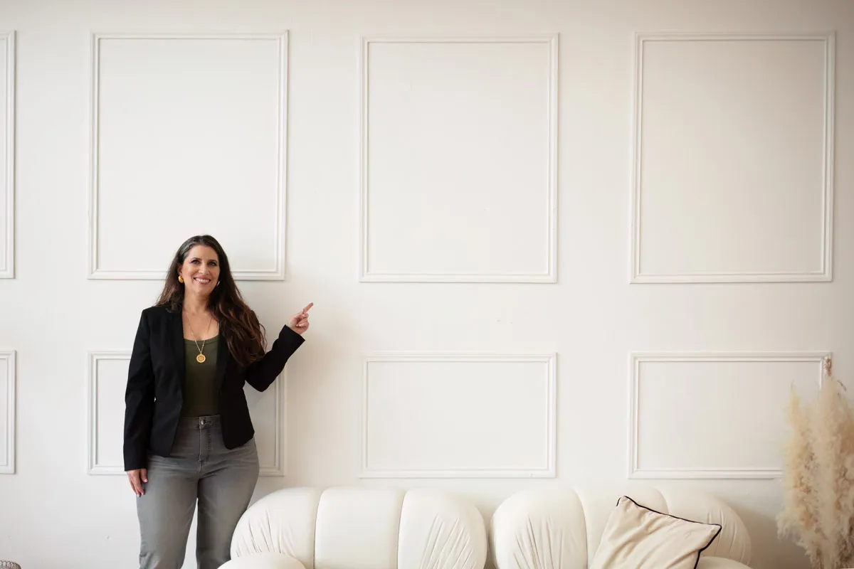 TJP_2024_04_26_0051 A woman in a black blazer and gray pants smiles and points at a blank decorative panel on a white wall above a white sofa, presenting her role as a wellness coach.
