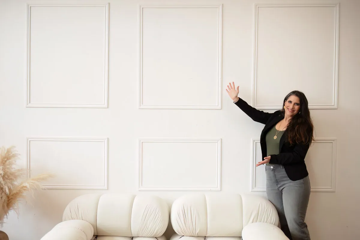 TJP_2024_04_26_0059 A wellness coach with long hair gesturing towards blank picture frames on a decorative wall above a white couch.
