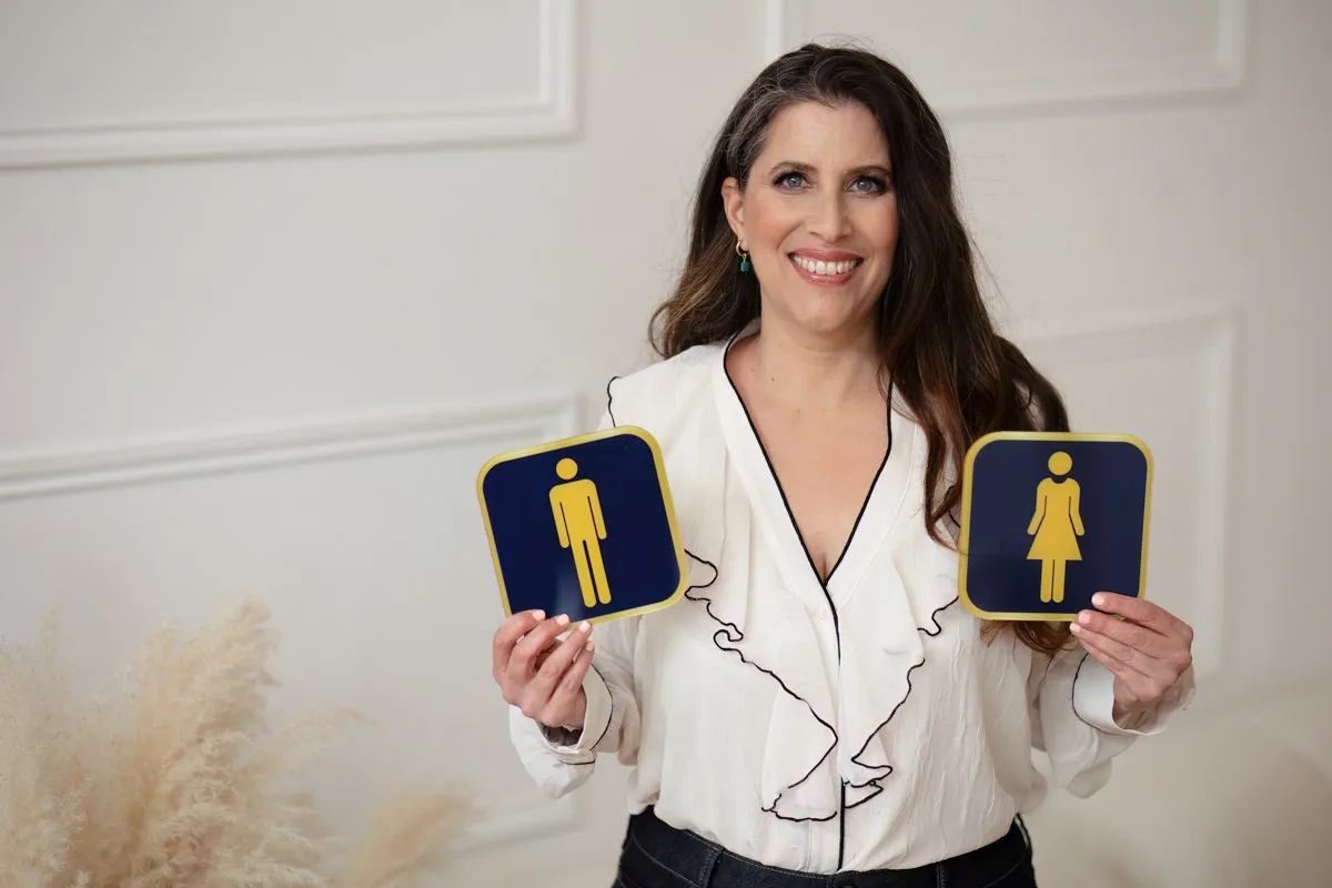 TJP_2024_04_26_0062 A smiling wellness coach holding up two restroom signs, one for males and one for females, against a white paneled background. She wears a white blouse with ruffle details.