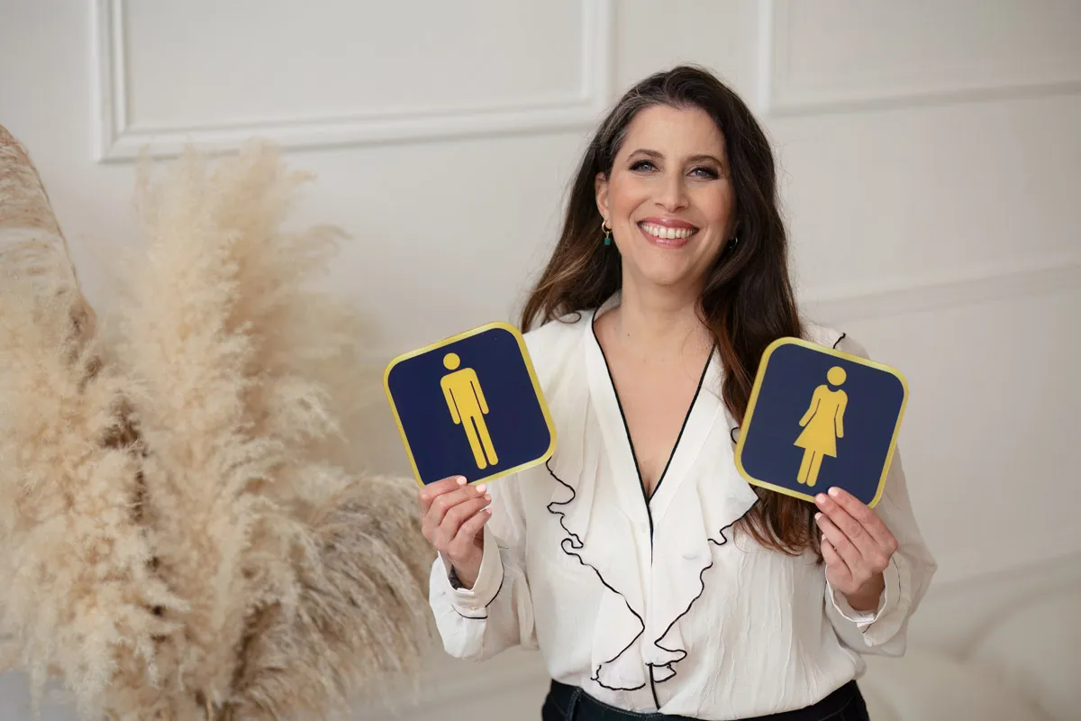 TJP_2024_04_26_0063 A smiling wellness coach in a white blouse holds up two restroom sign placards, one with a male icon and one with a female icon, in a well-lit room with soft background decor.