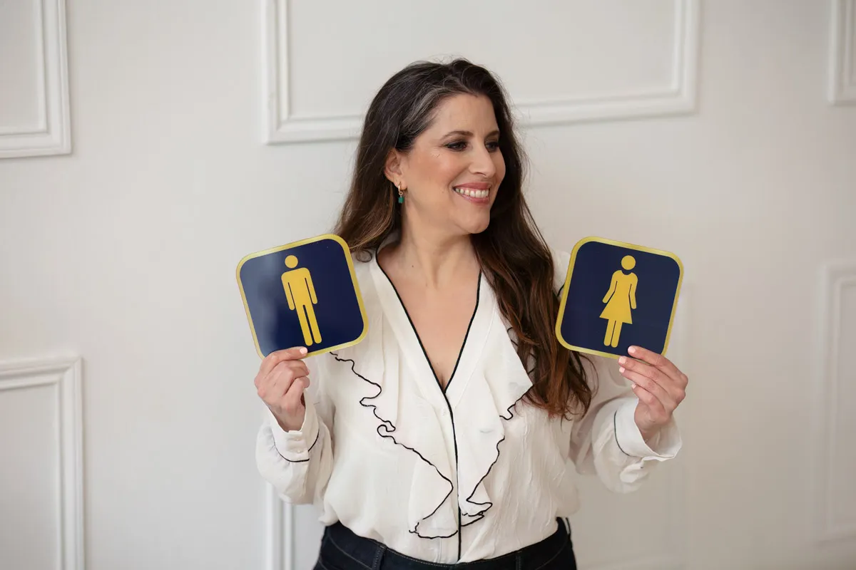 TJP_2024_04_26_0067 A wellness coach with long brown hair, wearing a white blouse, smiles as she holds up two restroom signs, one for men and one for women, against a white paneled wall.