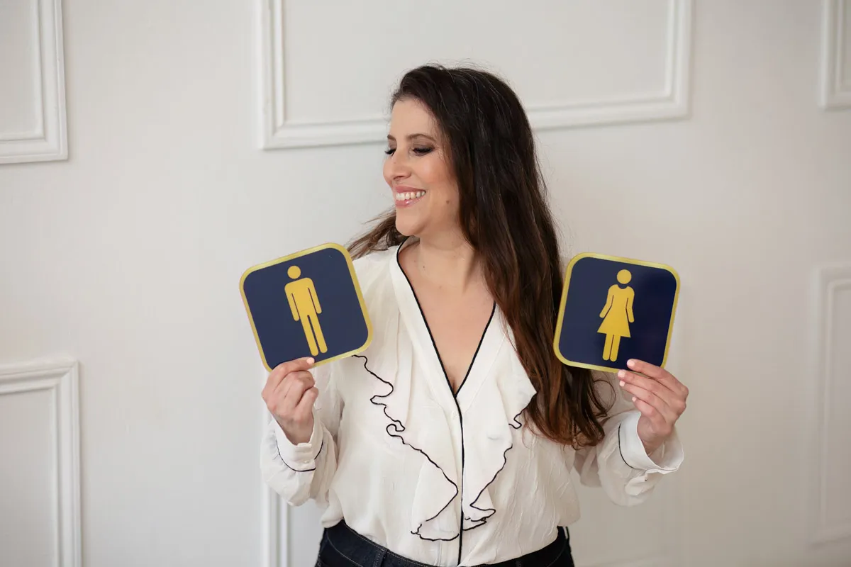 TJP_2024_04_26_0068 A smiling wellness coach holds up two restroom signs, one featuring a male icon and the other a female icon, against a plain white background.