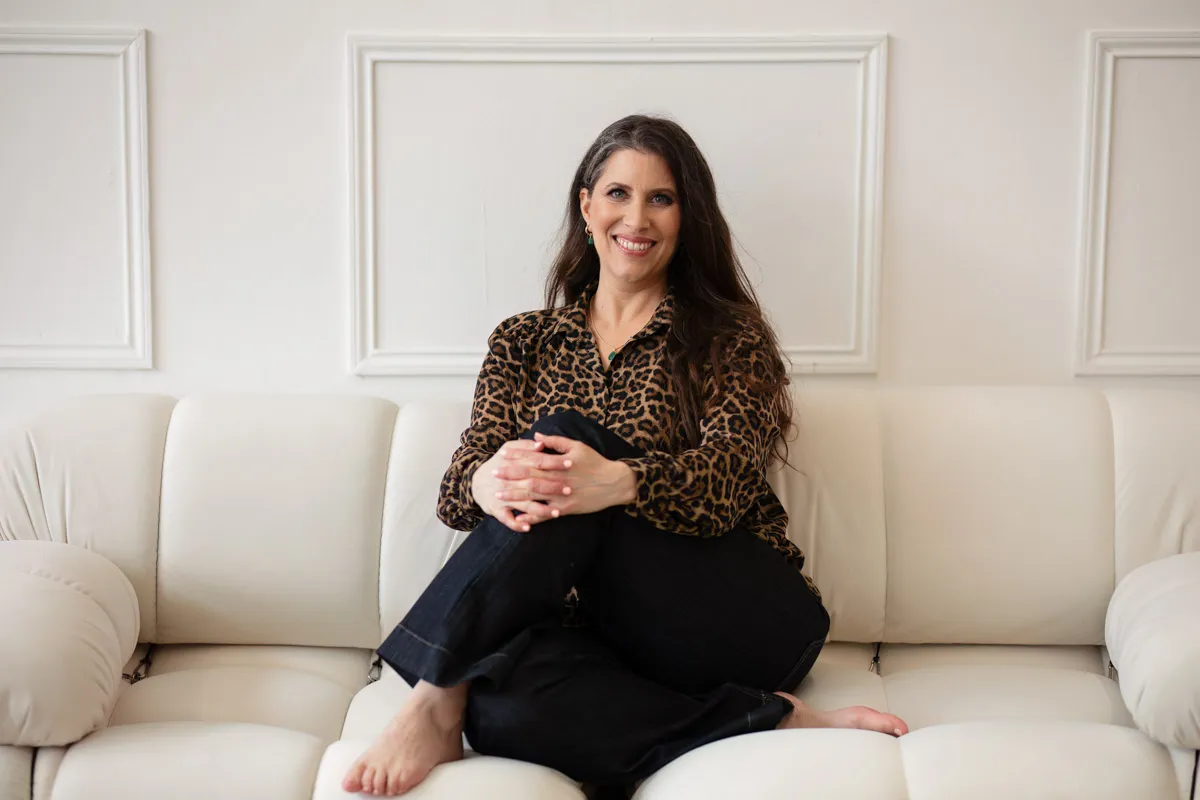 TJP_2024_04_26_0081 A woman with long, dark hair, wearing a leopard print blouse and black pants, sits barefoot on a white couch, smiling at the camera for a brand photography shoot.