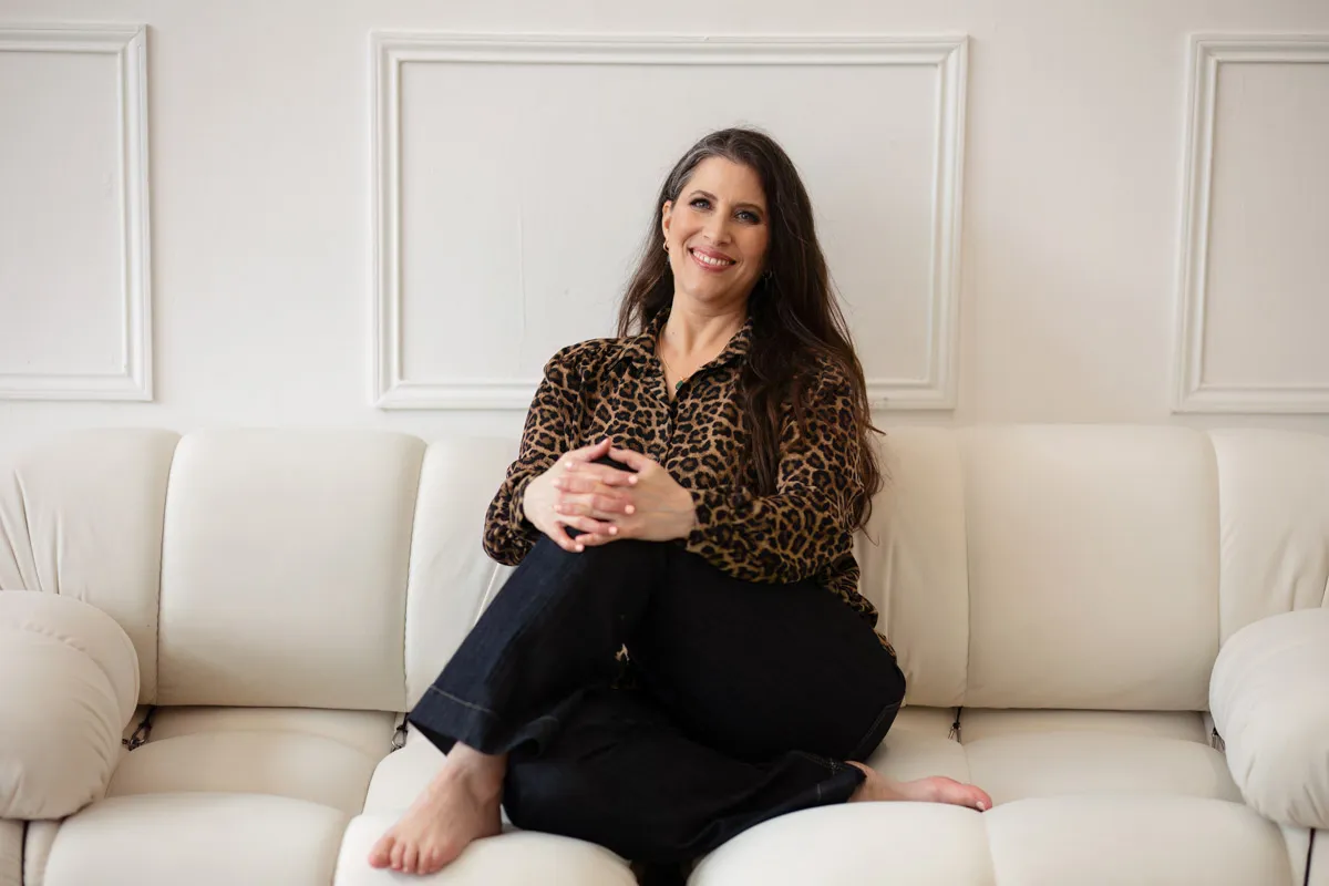 TJP_2024_04_26_0083 A woman with long dark hair, wearing a leopard print blouse and black pants, sits comfortably on a white sofa, smiling gently at the camera for her brand photography session.