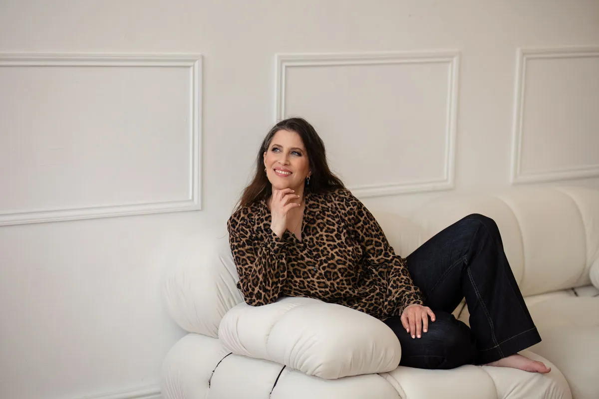 TJP_2024_04_26_0091 A woman in a leopard print blouse and jeans sits relaxed on a white couch, smiling and resting her chin on her hand in a bright, elegant room designed for brand photography.