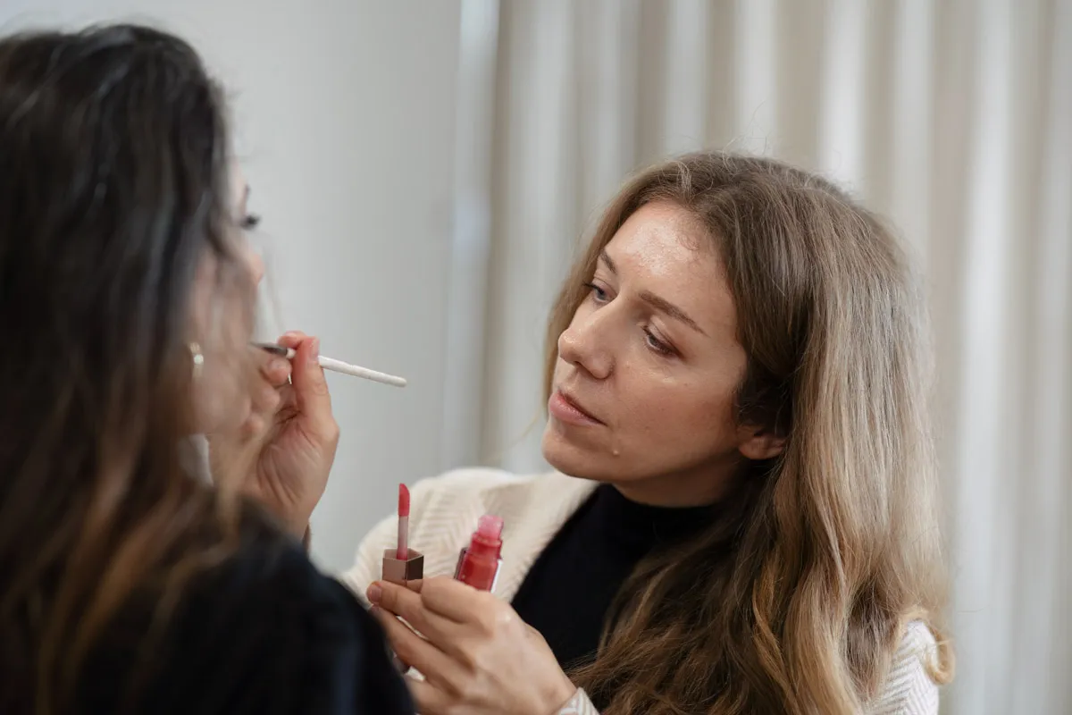 TJP_2024_04_26_0107 A woman in a black turtleneck concentrates as she applies makeup to another woman's face during a brand photography session, holding a lip product and a brush.