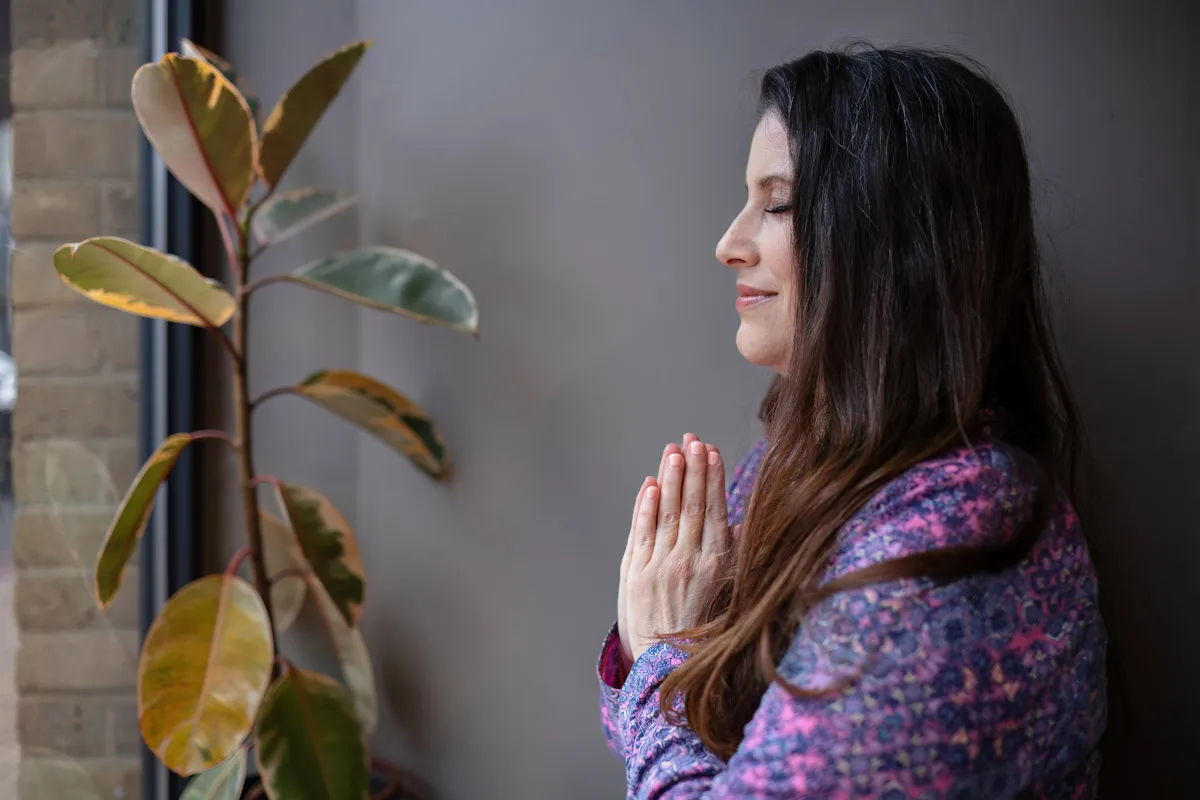 TJP_2024_04_26_0146 A woman with long hair, wearing a floral blouse, stands by a window with closed eyes and hands pressed together in a prayer-like gesture, next to a potted plant with large leaves, capturing the