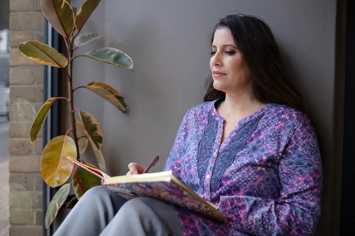 TJP_2024_04_26_0150 A woman sits by a window, holding a notebook and pen, deep in thought. She wears a patterned blouse and is surrounded by the peaceful ambiance of her wellness coach office enhanced by a potted