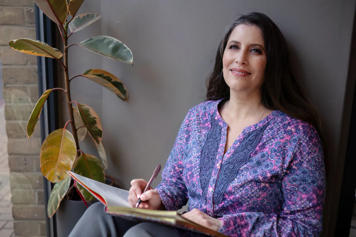 TJP_2024_04_26_0151 A woman sitting beside a window, holding a pen and notebook, smiles gently while wearing a paisley-patterned blouse. A potted plant is partially visible next to her. This serene setup enhances her