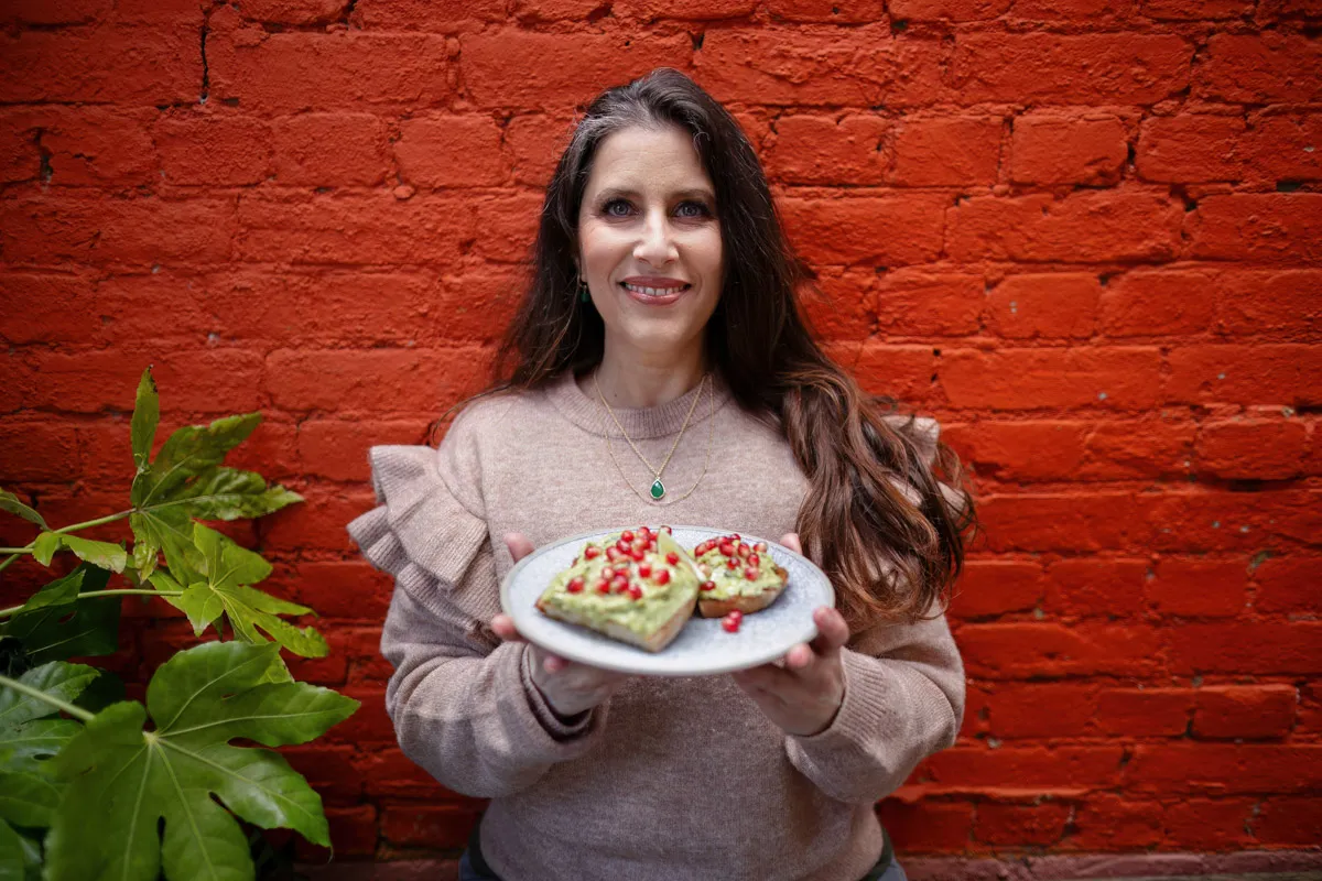 TJP_2024_04_26_0181 A woman in a beige sweater smiles while offering a plate with avocado toast garnished with pomegranate seeds, standing in front of a red brick wall with green plants beside her, perfectly capturing the
