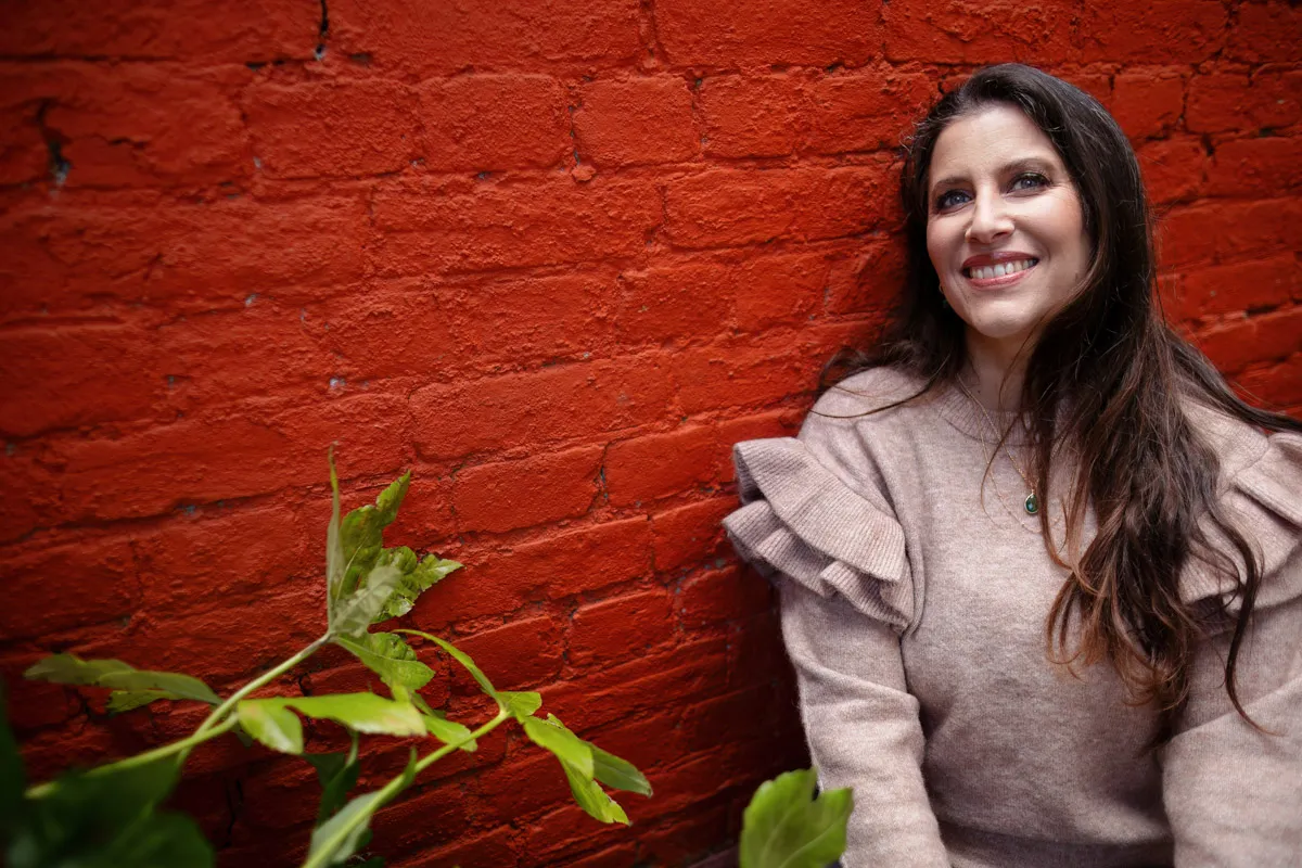 TJP_2024_04_26_0188 A smiling woman with long dark hair, wearing a grey sweater, sits in front of a vibrant red brick wall, holding a green leafy plant to her left, in a wellness coach brand photography session