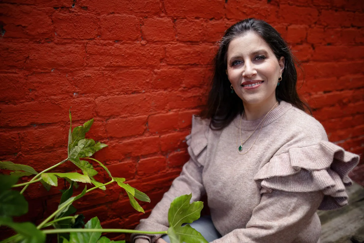 TJP_2024_04_26_0192 A woman smiling at the camera while sitting in front of a red brick wall, wearing a ruffled light brown sweater and a green pendant necklace, with green plants nearby. This setting illustrates her role as a Wellness Coach