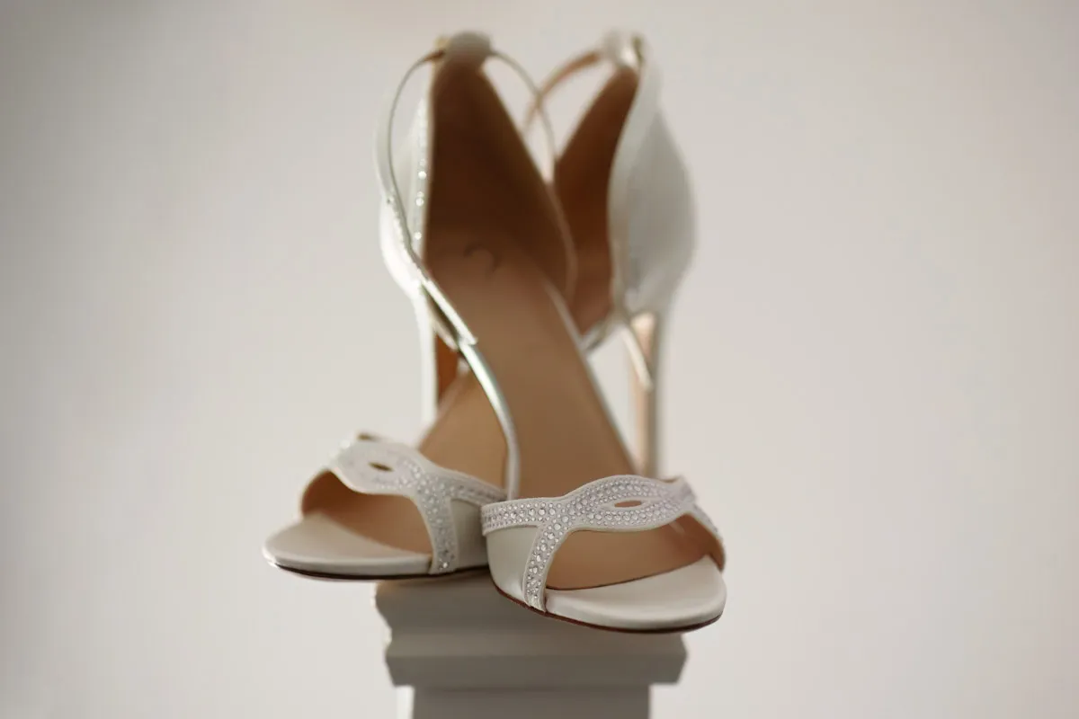 TJP_2023_06_24_0064 A pair of elegant white high-heeled sandals with open toes and sparkling embellishments on the straps, perfect for marrying your best friend, placed on a small white pedestal against a plain light-colored background.
